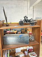 Full shelf view showing all four items: metal feather sculpture, welded motorcycle, galvanized box, glass vase with shells