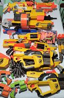 Top view showing multiple yellow blasters, drum magazine, blue blaster, small pistols, belt links and darts