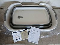 Collapsed white and grey baby bathtub with booklet and card on plastic wrap