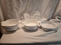 Three CorningWare Blue Cornflower casserole dishes side by side, two clear glass lids behind