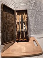 Open presentation box showing carving knife, fork, sharpening steel lined in tan felt alongside wood carving board