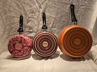 Three pans displayed bottom side up showing decorative patterns in pink floral, maroon rings, orange rings