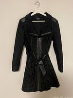 Full view of black floral jacquard Bebe jacket hanging on hanger showing front zipper, belt, faux leather accents