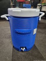 Front view of blue Rubbermaid drinking water cooler with lid, handles and spigot visible