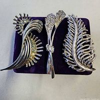 Three silver tone brooches on purple pad, front view
