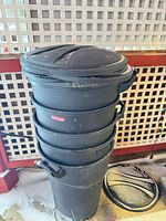 Stack of six black Rubbermaid garbage bins with lids, showing size and wear