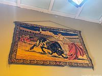 Full view of tapestry on wall showing bull, matador, arena, decorative border