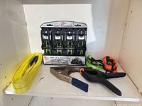 Shelf with packaged ratchet straps, yellow strap, hatchet/pry bar, black clamp tool, two loose straps partially visible