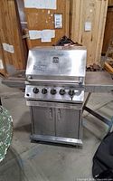 Front view of stainless steel gas grill showing hood, control panel with five knobs, side shelves and cabinet base