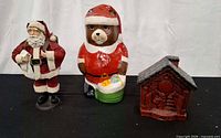 Front view of all three Christmas figurines: fabric Santa, ceramic bear Santa music box, red house decoration