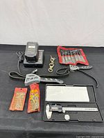 Overall view showing caliper case open with tool, battery charger with battery, chisel/punch roll, hex key sets, swivel hooks and hardware packs on black backdrop