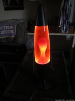 Lamp lit, orange-red wax rising inside glass vessel, black base visible