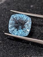 Cushion fantasy cut blue topaz in tweezers close-up
