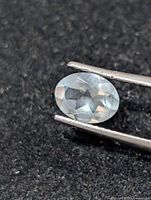 Oval faceted light blue gemstone held by tweezers against dark background