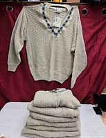 Stack of seven beige cable-knit sweaters with one displayed above