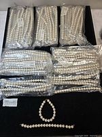 Multiple sealed dozen-count bags of faux pearl strands displayed on black surface