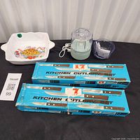 Group shot of all items: two blue cutlery set boxes, knives, USB chopper with cable, plastic bowl, ceramic pasta dish