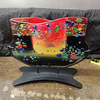 Front view of fused glass sculpture on stand showing wave shape and multicolored nuggets