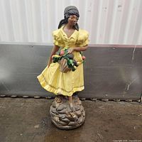Front view of ceramic statue showing woman, dress color, basket contents, and rocky base