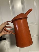 Side view of orange metal oil can showing handle, spout, overall form, and paint wear