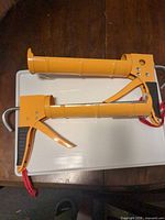 Two unused yellow caulking guns on tray showing full view of barrel, trigger, grip and lanyard