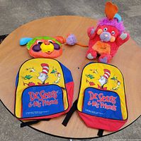 Group shot of Popples coin bank plush, Popples mask, two Dr. Seuss backpacks on round table