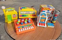 All six toys arranged outdoors on table including Sesame Street playset, Grand Piano, Big Mouth Singers, Talking Tutor Robot with box, kaleidoscope, cards