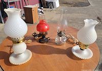 Group shot of two white hobnail electric lamps, red glass electric lamp, and clear glass oil lamp on table