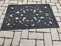 Full view of black rubber doormat on paving stones showing decorative cut-out pattern and overall condition
