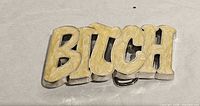 Front view of silver-tone metal belt buckle with cream marbled letters forming the word “Bitch”