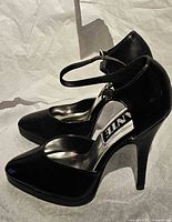 Side view of both black patent high heel shoes showing ankle strap and heel profile