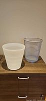 White plastic and silver mesh wastebaskets side by side on wooden surface