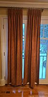 Pair of rust-red jacquard curtain panels hanging on rod showing full length and pinch pleats