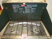 Overall view of open Coleman three-burner propane stove showing lid, burners, grate, and instruction decal