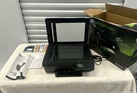 Printer with lid open showing flatbed scanner, accessories and documents beside, original box to the right