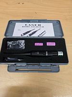 Closed gray plastic case opened to show full contents: laser bore sighter, adapter bag, two pink CR2 batteries, small tool, instruction card.