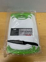 Sealed package containing three nested green-edged white cutting boards and separate black chef knife