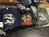 Three unfolded T-shirts (navy, black, olive) plus stacks of bagged shirts