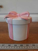 Side view of ceramic gift box jar with ruler for scale showing pink stripes and small chip on ribbon