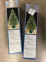 Two boxed 32 inch fiber optic Christmas trees standing upright on a tiled floor. Boxes show product images, instructions, and branding for fiber optic Christmas trees with green trees and gold decorations.