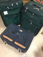 Set of three suitcases placed on the floor, two large dark green fabric with multiple zippered compartments, and one smaller navy blue suitcase with leather accents.
