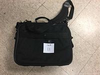 Photo of a black computer bag lying flat on a tiled floor with a small paper labeled 'lot 11' placed on it.