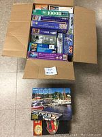 Open cardboard box on tiled floor filled with multiple boxed puzzles including 1000-piece and 500-piece puzzles, showing various scenic images.