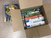 Photo showing an open cardboard box filled with multiple boxed jigsaw puzzles with visible piece counts 500, 750, and 1000 pieces. Puzzle boxes from multiple brands are stacked tightly in the box.