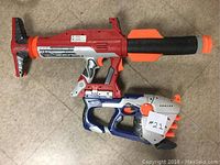 Two large Nerf guns on the floor: one red, silver, and black with orange tip; the other blue, white, and orange.