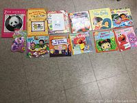 Photo of 12 children's books arranged in a row on a floor, showing colorful covers and varied themes such as animals, family stories, nursery rhymes, and characters like Elmo.
