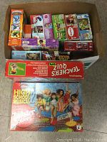Top-down view of a box containing multiple boxed games and puzzles with visible titles including High School Musical Twister Moves and Teacher's Quiz.