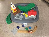Gray plastic container with green curved track pieces, a lighthouse, a red-roofed building, yellow crane building, and small blue and red train cars arranged on top.