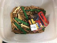 Bin containing light brown and green wooden building logs with a small clear bag filled with colorful plastic accessories used for construction play.