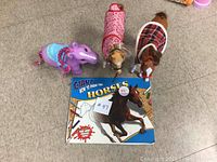Four items on floor including two toy horses, a purple pony toy, and a drawing book about horses.